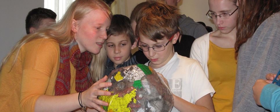 Mehrere Kinder und Jugendliche betrachten gemeinsam einen Globus. Sie zeigen interessiert auf verschiedene Länder und stehen dicht beieinander in einem hellen Raum.