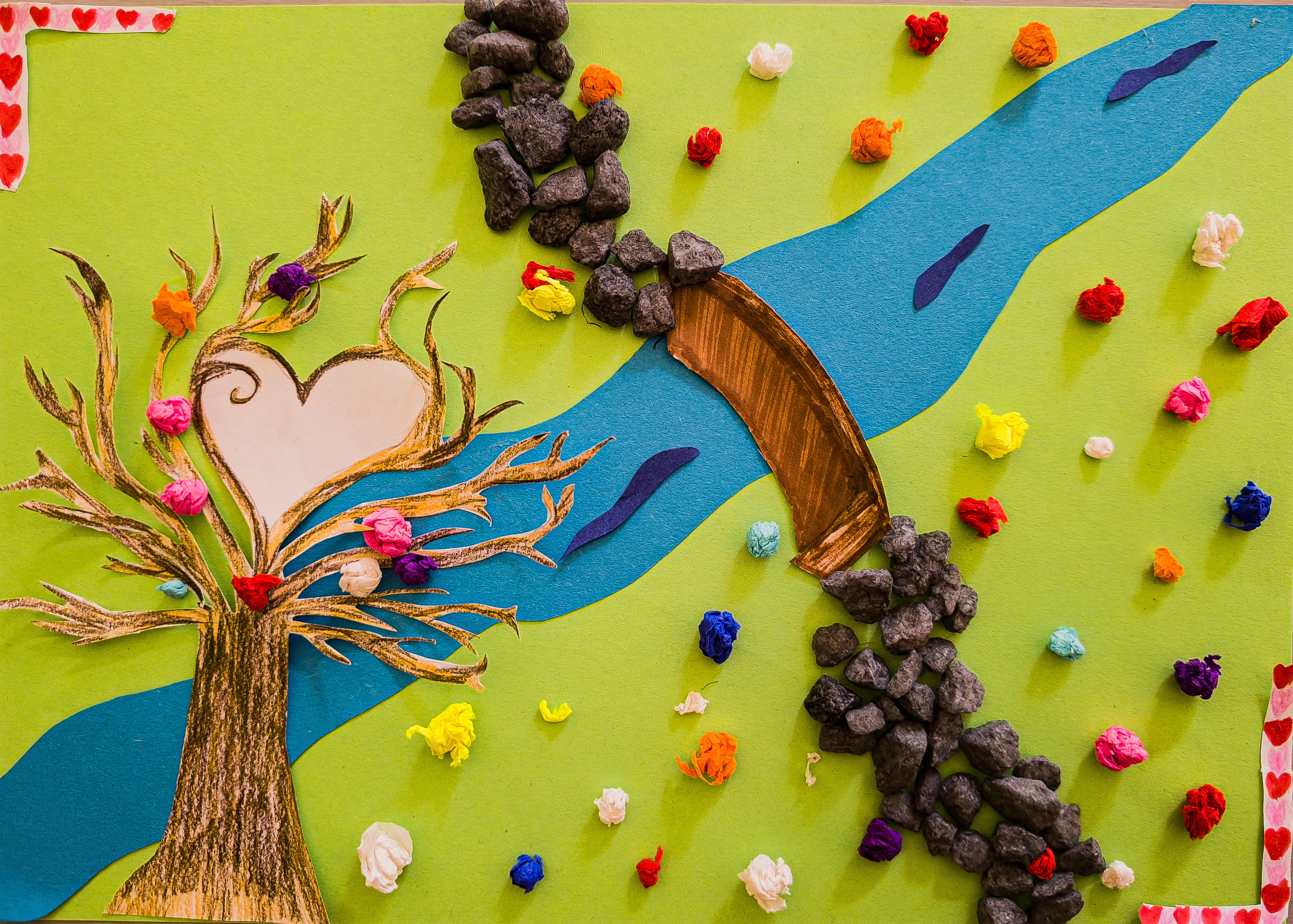 Ein buntes, gebasteltes Bild zeigt einen blauen Fluss, der sich durch eine grüne Landschaft zieht. Auf beiden Seiten des Flusses liegen schwarze Steine, eine braune Brücke führt darüber. Links steht ein goldbrauner Baum mit einem weißen Herz in der Krone, aus dem bunte Papierblumen wachsen. Über das ganze Bild verteilt sind weitere kleine, bunte Papierkügelchen.